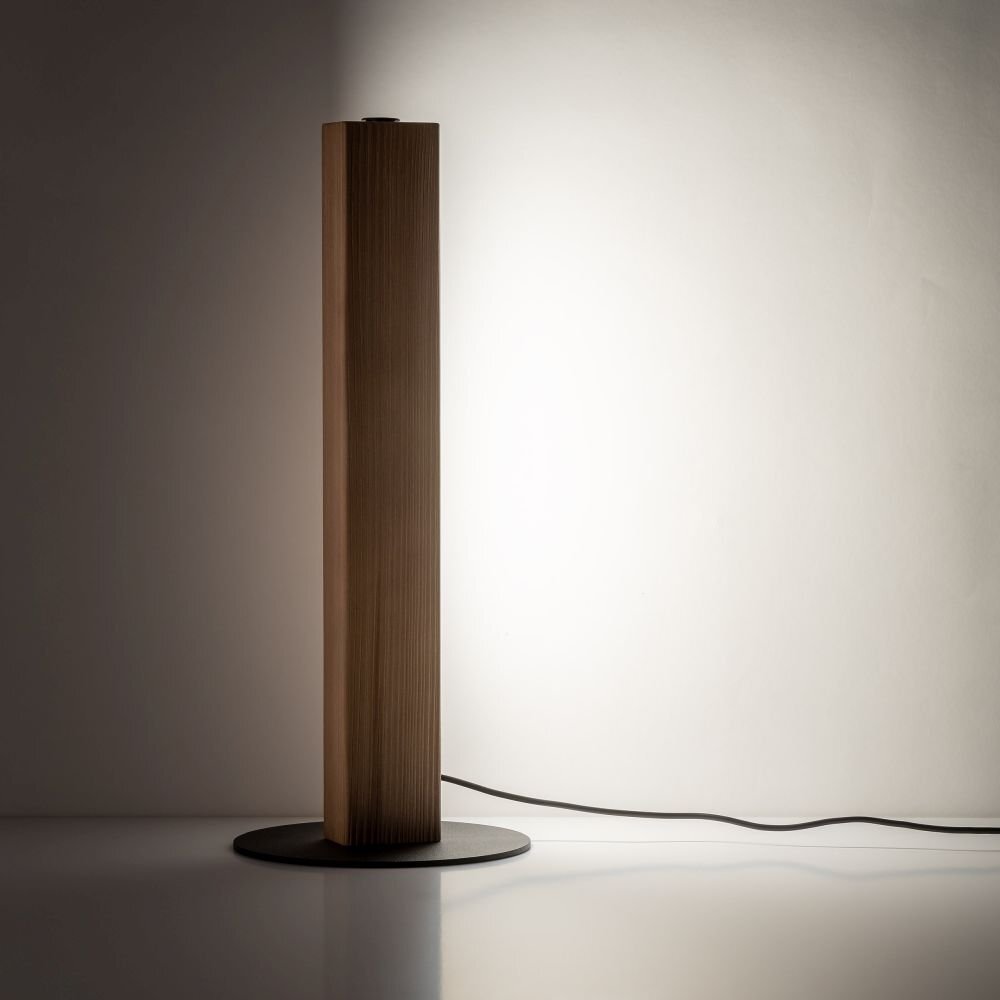 Na obrazie widoczna jest lampa stołowa o prostokątnym kształcie, wykonana z drewna w kolorze brązowym, stojąca na okrągłej podstawie. Lampa emituje ciepłe światło, a w tle widać jej odbicie na ścianie.