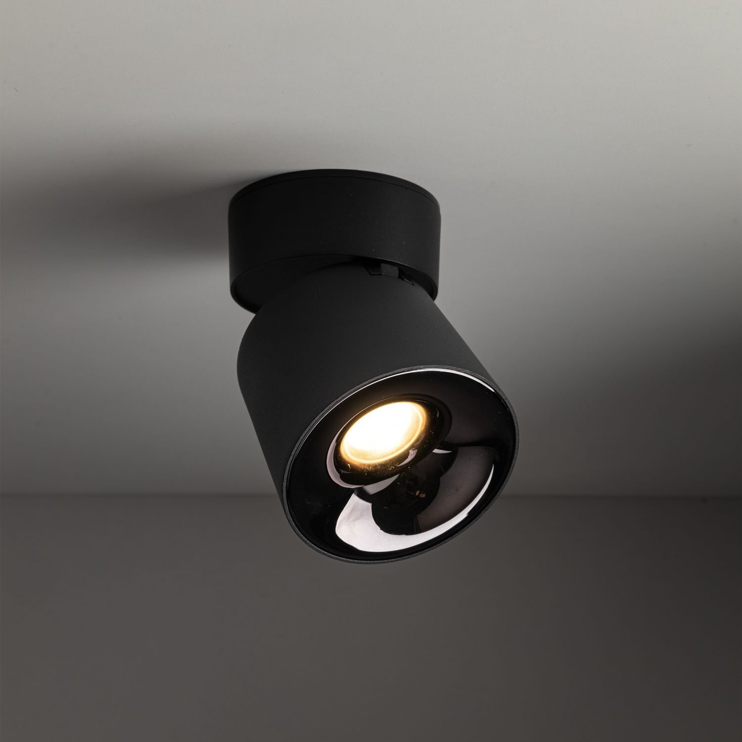 Czarna lampa reflektorowa LED zamontowana na suficie, emitująca ciepłe białe światło. Lampa ma cylindryczny kształt i jest ruchoma.