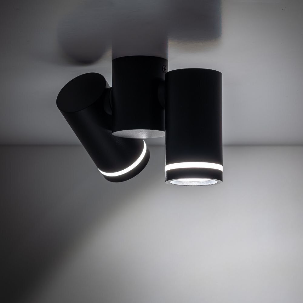 Czarna lampa sufitowa z dwoma regulowanymi reflektorami w kształcie cylindrów, zamontowana na suficie. Reflektory emitują delikatne światło wokół krawędzi.