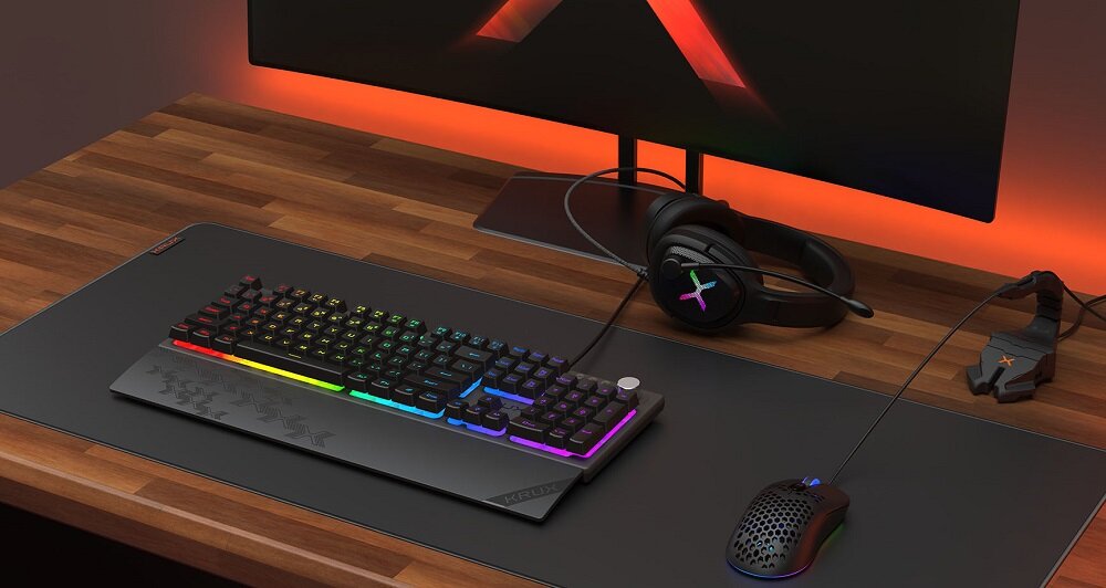 Na drewnianym biurku znajduje się klawiatura z podświetleniem RGB, myszka oraz słuchawki. W tle widoczny jest monitor z logo \'X\'.