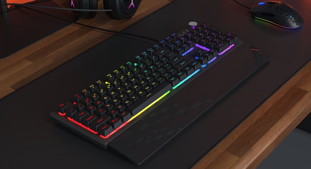 Na biurku znajduje się klawiatura z podświetleniem RGB, obok której leżą słuchawki i myszka. Klawiatura ma ciemną obudowę i kolorowe podświetlenie klawiszy.