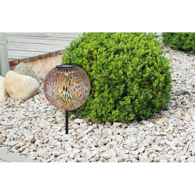 Lampa solarna LED o ażurowym wzorze umieszczona na kamiennym podłożu obok zielonego krzewu. W tle widoczny jest drewniany taras i kilka dużych kamieni.