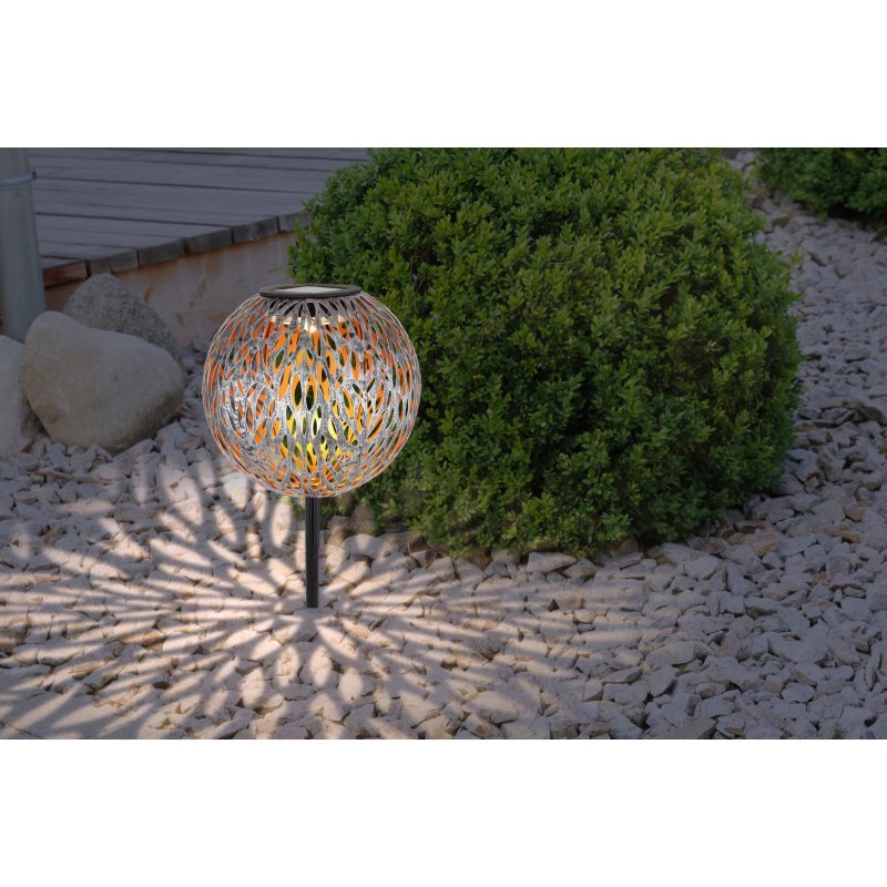 Lampa solarna LED o ażurowym wzorze, emitująca światło na kamienistym podłożu, obok zielonego krzewu. W tle widoczna jest drewniana powierzchnia i duże kamienie.