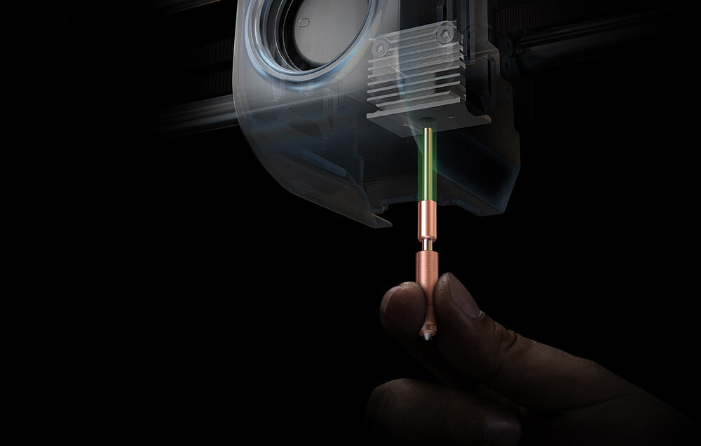 Obraz przedstawia rękę trzymającą miedziany element z zielonym prętem, który jest częścią mechanizmu drukarki 3D. W tle widoczny jest fragment urządzenia z metalowym radiatorem.