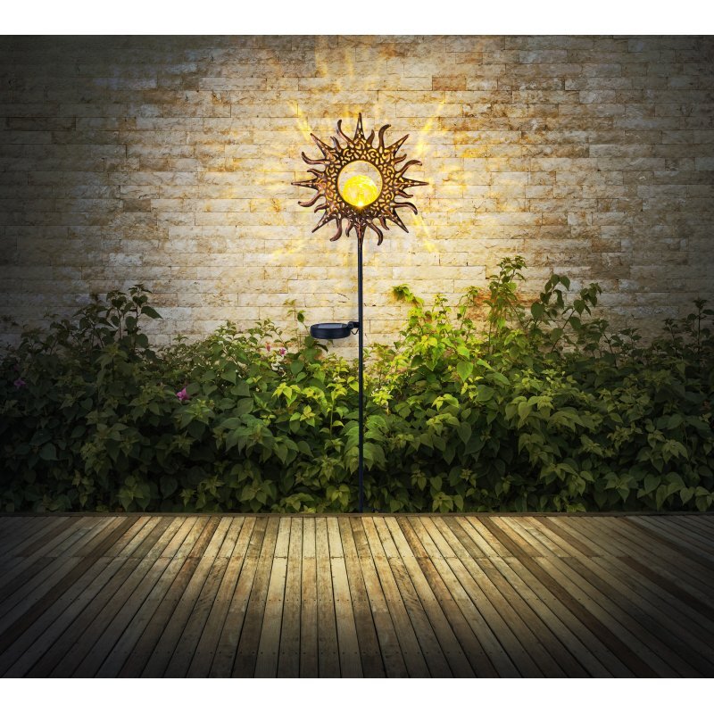 Lampa solarna w kształcie słońca oświetla przestrzeń przed ścianą z cegieł, otoczona zielonymi roślinami. Podłoga wykonana jest z drewnianych desek.