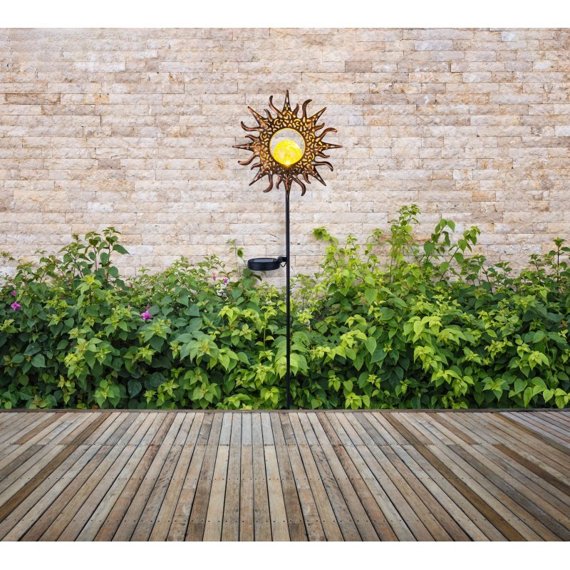 Lampa solarna w kształcie słońca z żółtym środkiem umieszczona jest na tle kamiennej ściany i zielonych krzewów. Przed lampą znajduje się drewniana podłoga tarasowa.