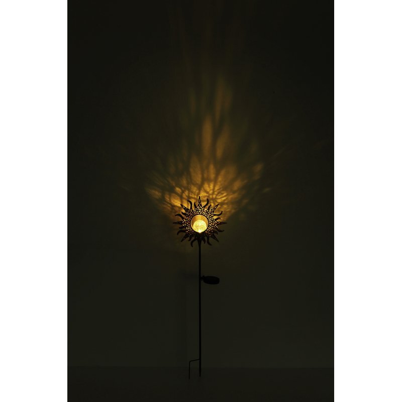 Lampa solarna w kształcie słońca emituje ciepłe, żółte światło, tworząc wzory na ciemnym tle. Lampa jest zamontowana na cienkim, metalowym pręcie.