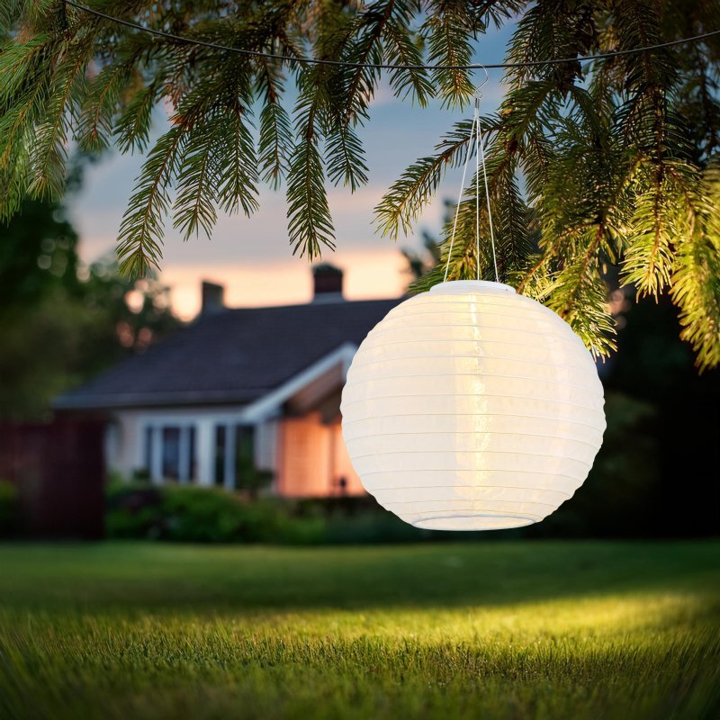 Na obrazie widoczna jest wisząca, biała lampa solarna w kształcie kuli, zawieszona na gałęzi drzewa. W tle znajduje się dom z ogrodem o zmierzchu.