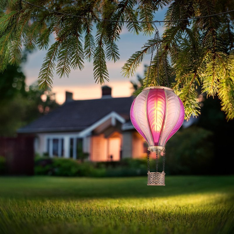 Na obrazie widać wiszącą lampę solarną w kształcie balonu, oświetloną i zawieszoną na gałęzi drzewa. W tle znajduje się dom z oświetlonymi oknami, otoczony zielonym trawnikiem.