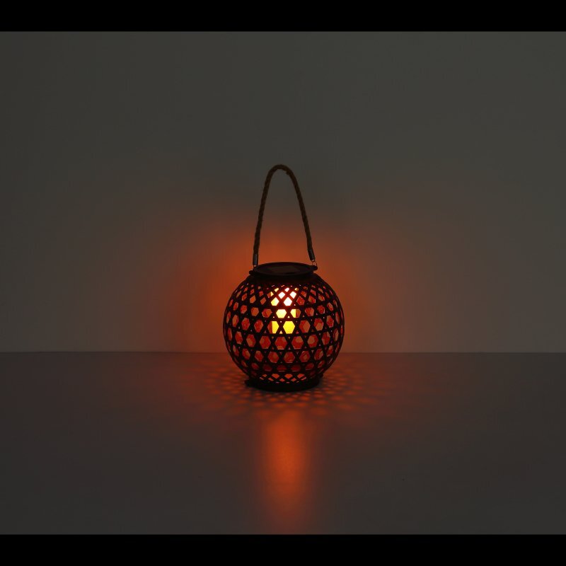 Na obrazie widoczna jest okrągła lampa solarna z ażurowym wzorem, emitująca ciepłe, pomarańczowe światło. Lampa posiada uchwyt do zawieszenia.