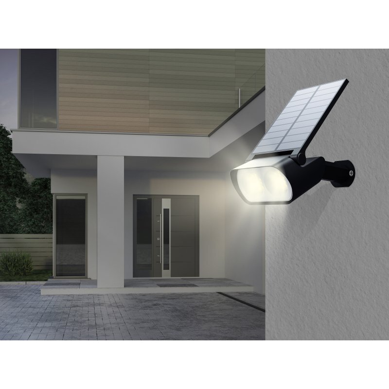 Na ścianie budynku zamontowana jest czarna lampa solarna z panelem słonecznym, emitująca jasne światło. W tle widoczny jest nowoczesny budynek z drzwiami wejściowymi i dużymi oknami.