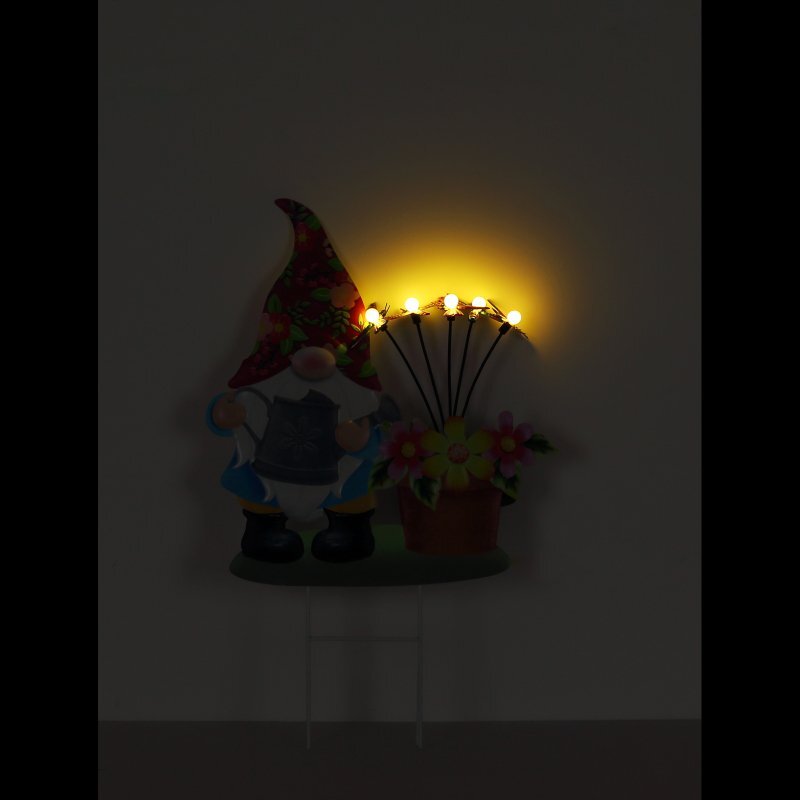 Na obrazie widoczna jest lampa solarna w kształcie krasnala z czerwonym kapeluszem, obok której znajduje się doniczka z kwiatami i świecącymi żółtymi diodami LED. Tło jest ciemne, co podkreśla światło emitowane przez diody.
