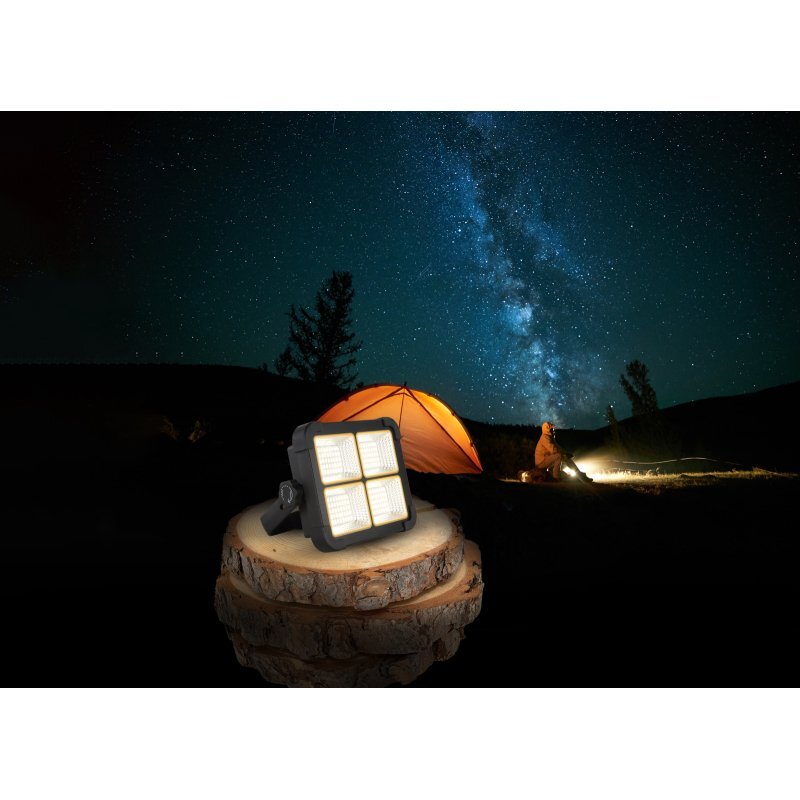 Na pierwszym planie widoczna jest lampa z czterema panelami LED, umieszczona na drewnianym krążku. W tle znajduje się rozgwieżdżone niebo, pomarańczowy namiot i osoba siedząca obok źródła światła.