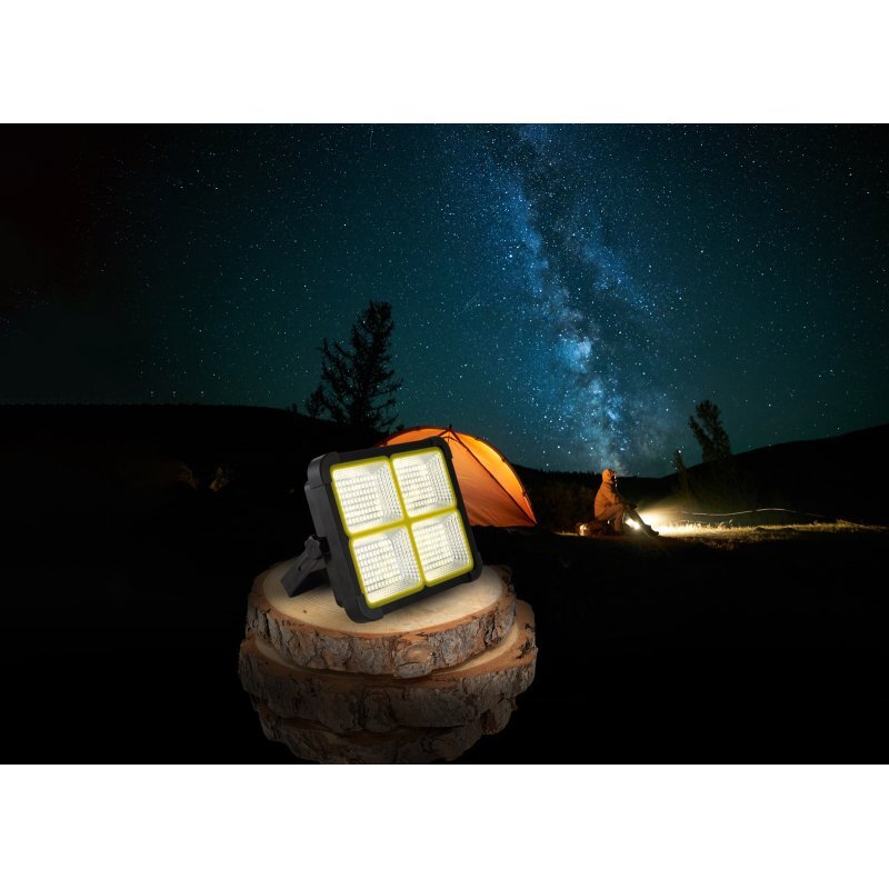 Na pierwszym planie widoczna jest lampa solarna LED z czterema panelami świetlnymi, ustawiona na drewnianym pniu. W tle znajduje się rozgwieżdżone niebo oraz pomarańczowy namiot oświetlony latarką.