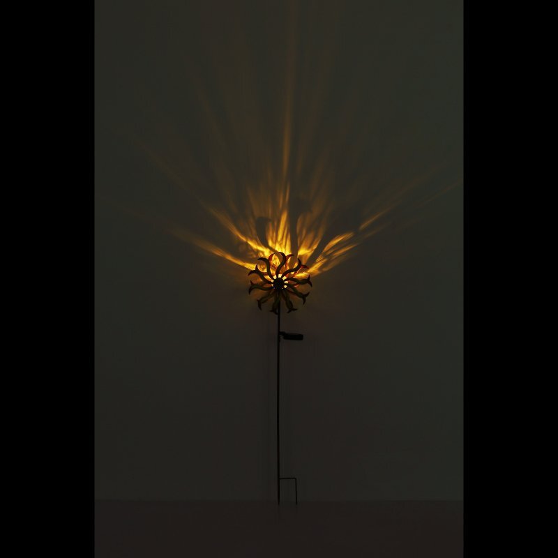 Na ciemnym tle widać lampę solarną z efektem świetlnym przypominającym promienie słońca. Lampa jest zamontowana na długim pręcie.