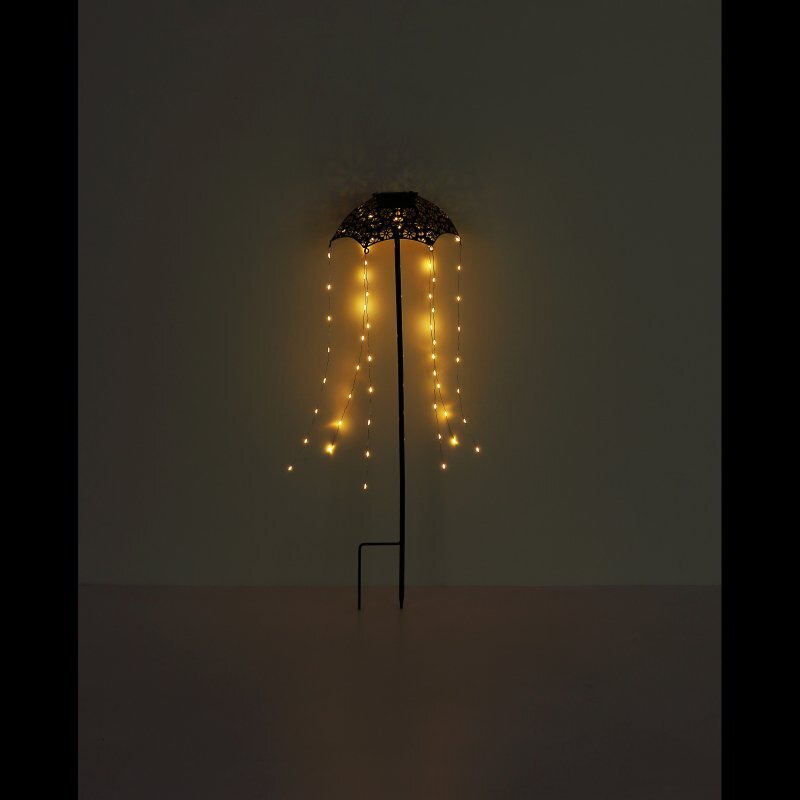 Na ciemnym tle widoczna jest lampa solarna w kształcie parasola z wiszącymi, świecącymi na żółto diodami LED. Lampa jest zamocowana na cienkim pręcie wbitym w podłoże.