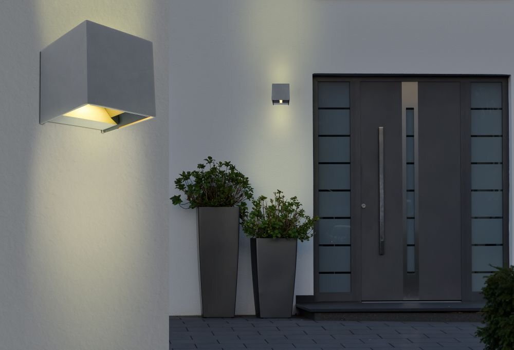 Na obrazie widoczna jest nowoczesna lampa ścienna zamontowana na zewnętrznej ścianie budynku, emitująca światło w górę i w dół. Obok znajdują się drzwi wejściowe oraz dwie wysokie donice z roślinami.