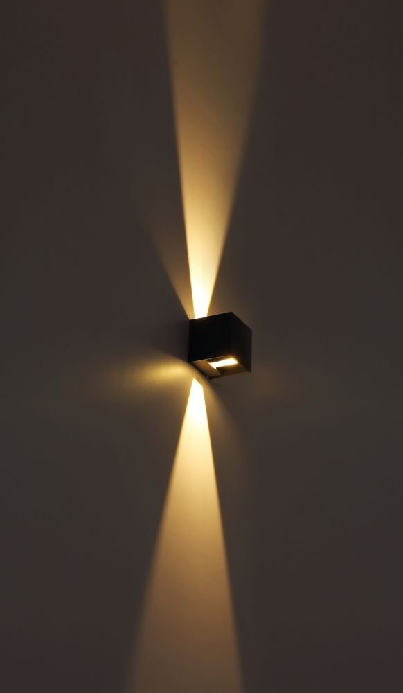 Na obrazie widoczna jest lampa ścienna emitująca światło w górę i w dół, tworząc efektowne snopy światła na ścianie. Lampa ma prostokątny kształt i ciemny kolor.