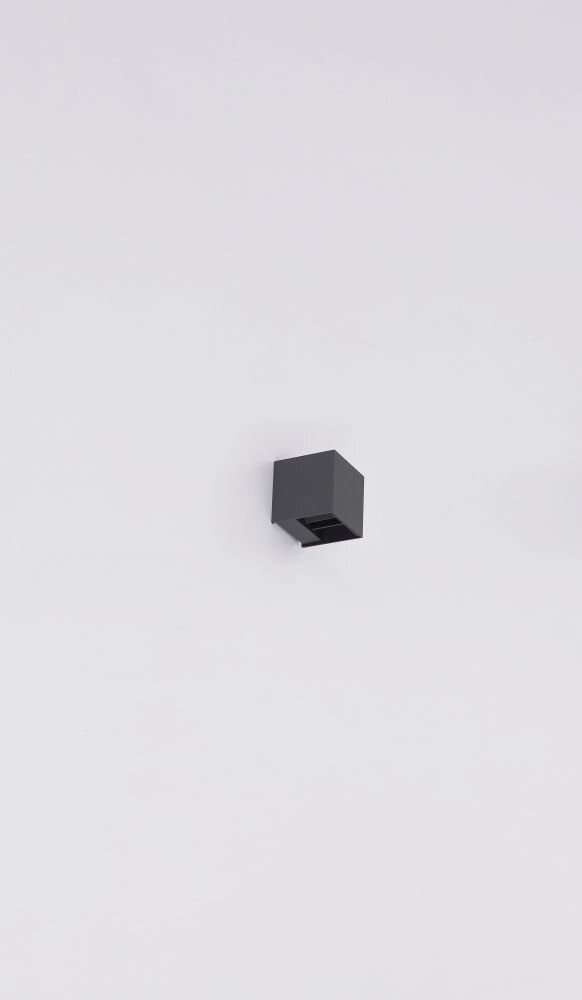 Na obrazie widoczna jest antracytowa lampa ścienna o prostokątnym kształcie zamontowana na jasnej ścianie. Lampa ma minimalistyczny design i skierowana jest w dół.