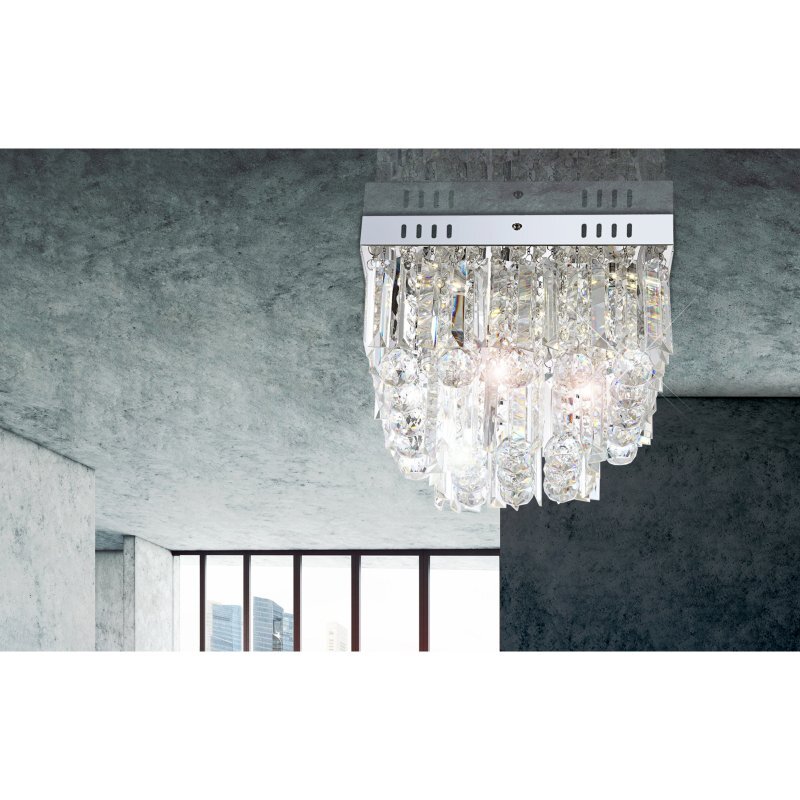 Lampa sufitowa z chromowanym wykończeniem i akrylowymi kryształami wisi na suficie w nowoczesnym wnętrzu. W tle widoczne są duże okna z widokiem na miasto.