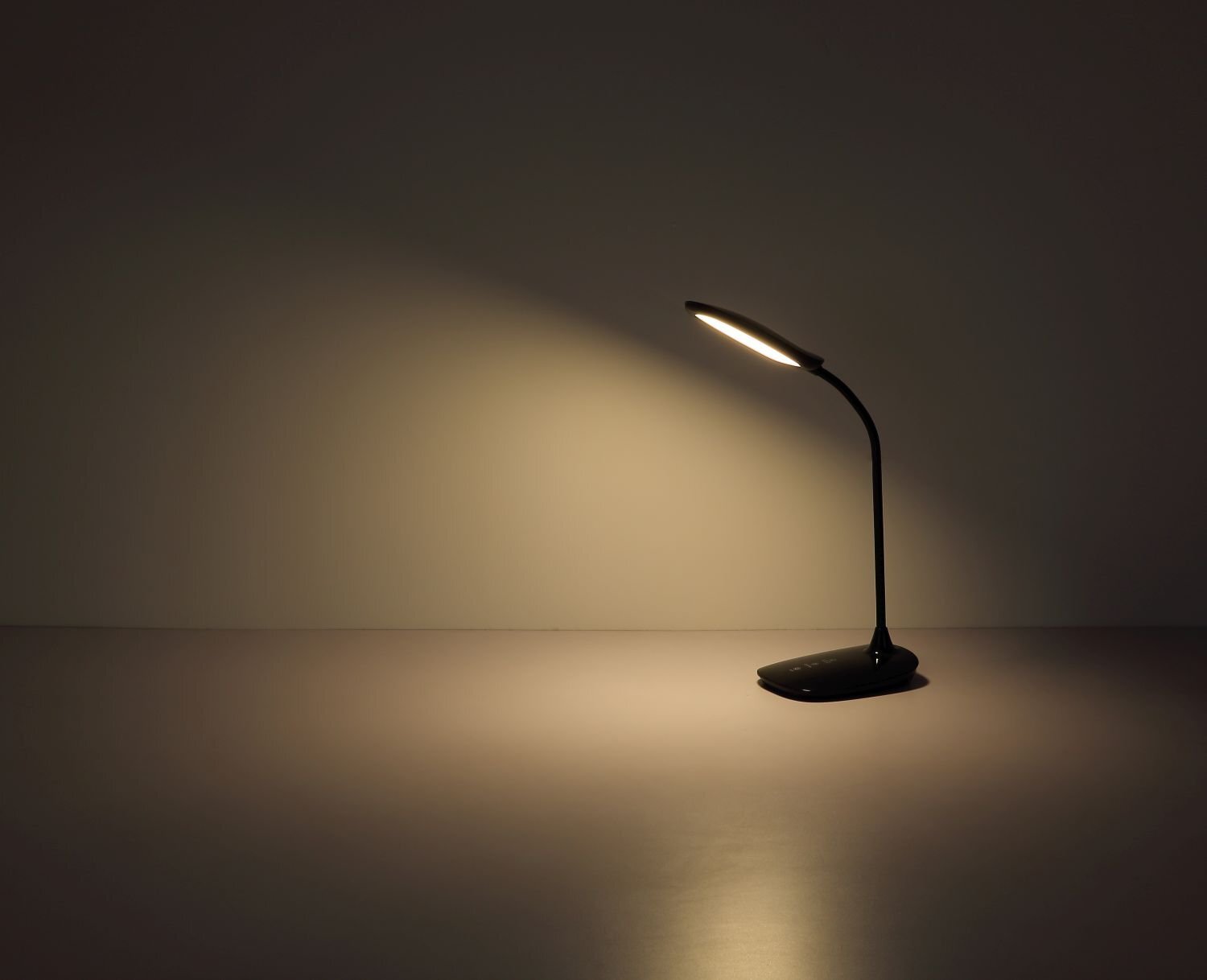 Na obrazie widoczna jest nowoczesna lampa biurkowa LED emitująca ciepłe światło na ciemnym tle. Lampa ma smukły, zakrzywiony kształt i stoi na płaskiej podstawie.