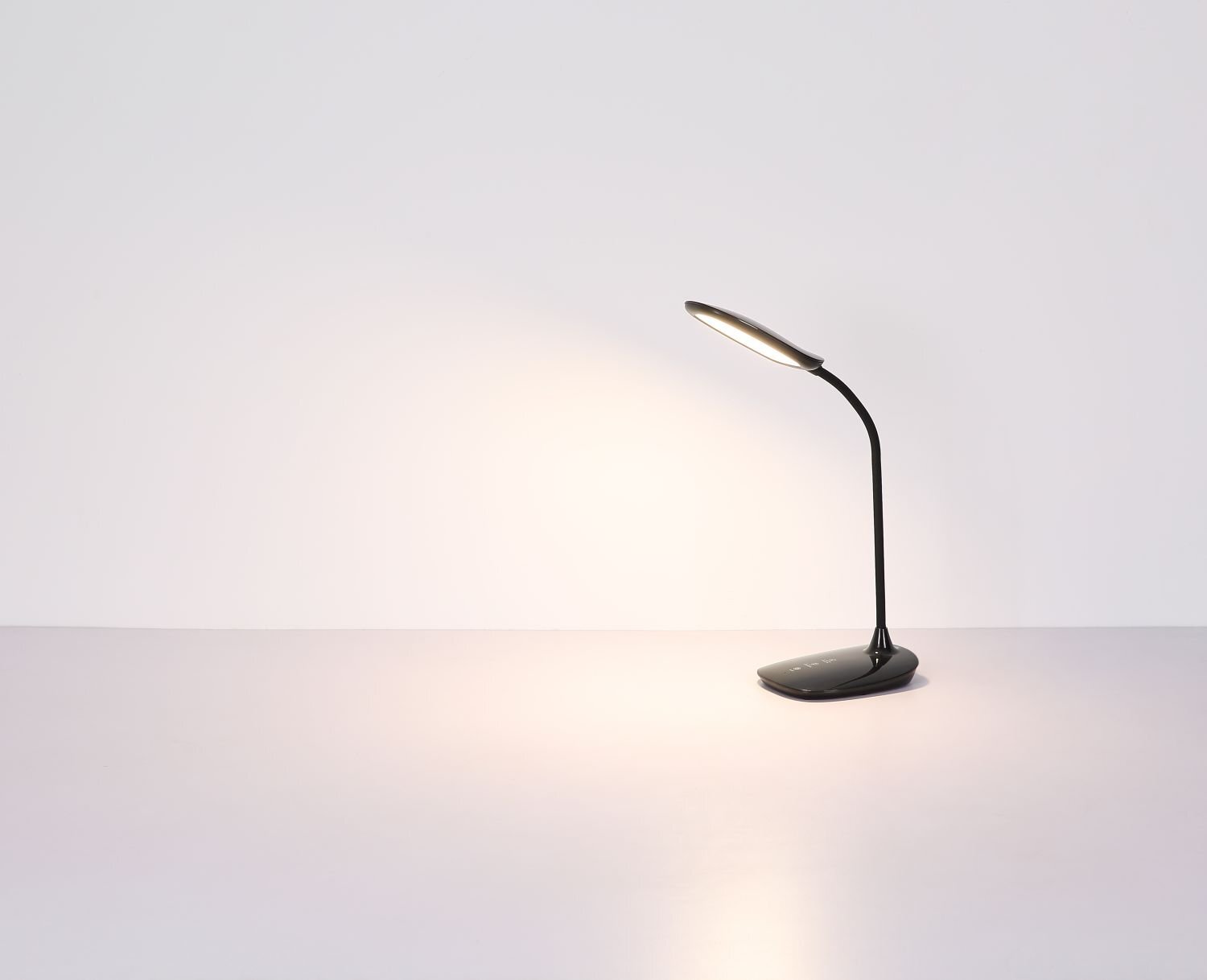 Na obrazie widoczna jest nowoczesna lampa biurkowa LED z czarnym, giętkim ramieniem i owalnym kloszem, emitująca ciepłe światło na jasnym tle.