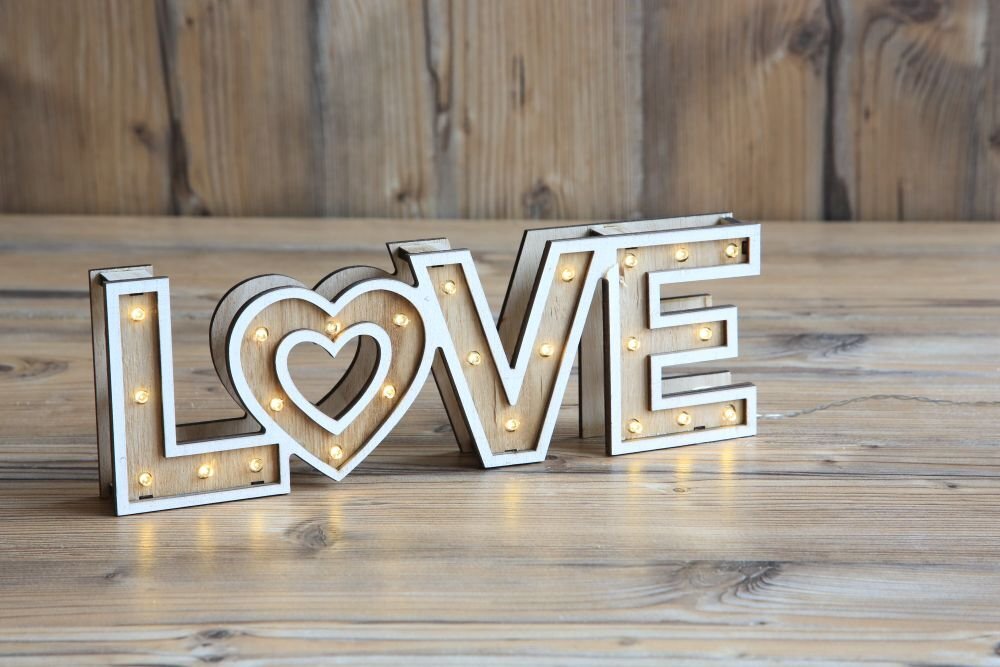 Drewniana lampa stołowa w kształcie napisu \'LOVE\' z wbudowanymi małymi żarówkami LED. Lampa stoi na drewnianym tle.