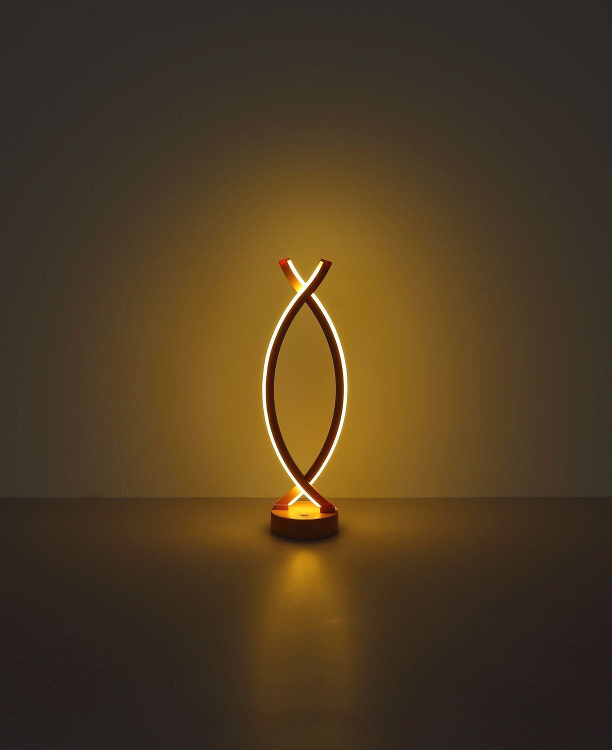 Nowoczesna lampa stołowa LED o kształcie dwóch skrzyżowanych łuków, emitująca ciepłe, żółte światło. Lampa stoi na okrągłej podstawie, a jej światło tworzy delikatną poświatę na powierzchni.