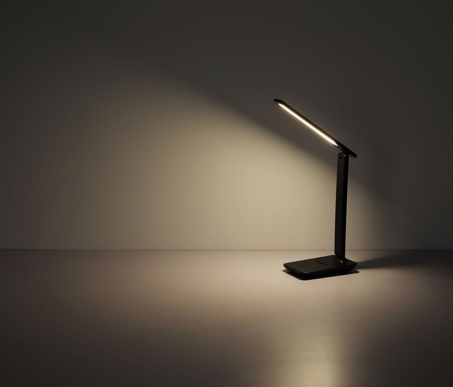 Na obrazie widoczna jest nowoczesna lampa biurkowa LED emitująca ciepłe światło na powierzchnię. Lampa ma smukły, minimalistyczny design i stoi na płaskiej podstawie.