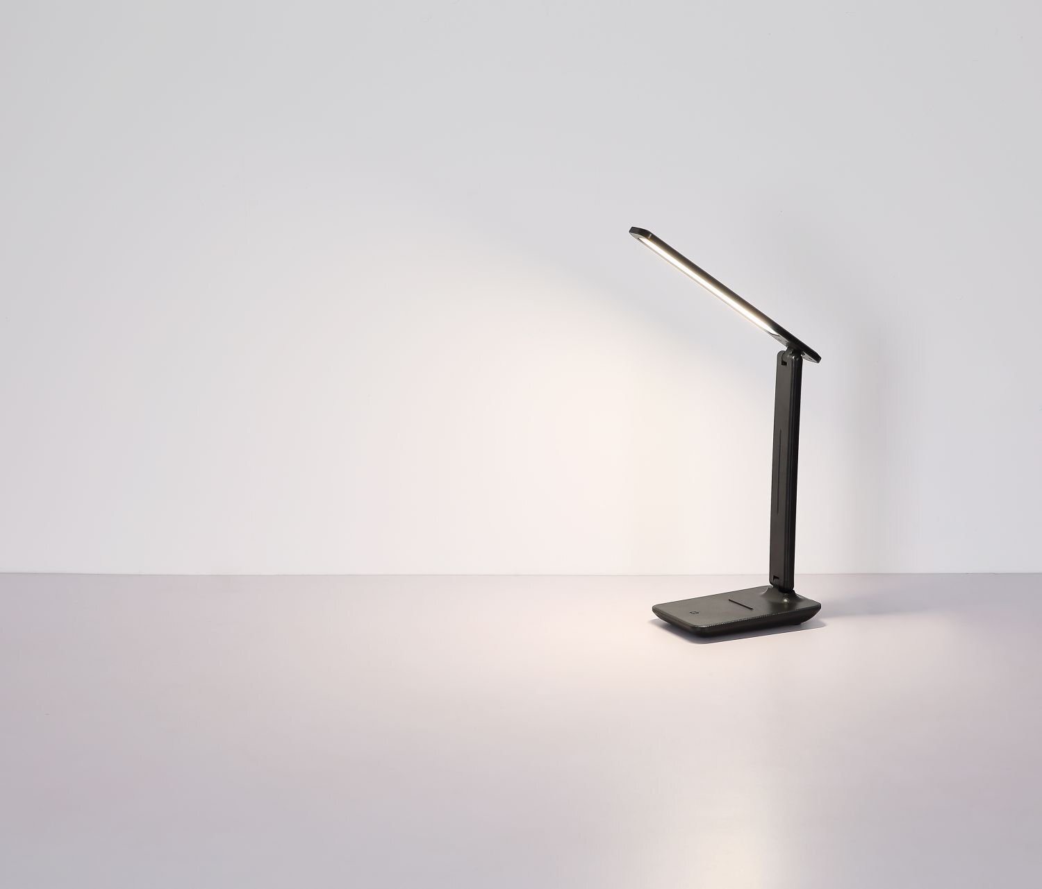 Na obrazie widoczna jest nowoczesna lampa biurkowa LED o prostym, minimalistycznym designie, emitująca jasne światło na powierzchnię stołu. Lampa ma ciemny kolor i składa się z prostokątnej podstawy oraz regulowanego ramienia z wąskim panelem świetlnym.