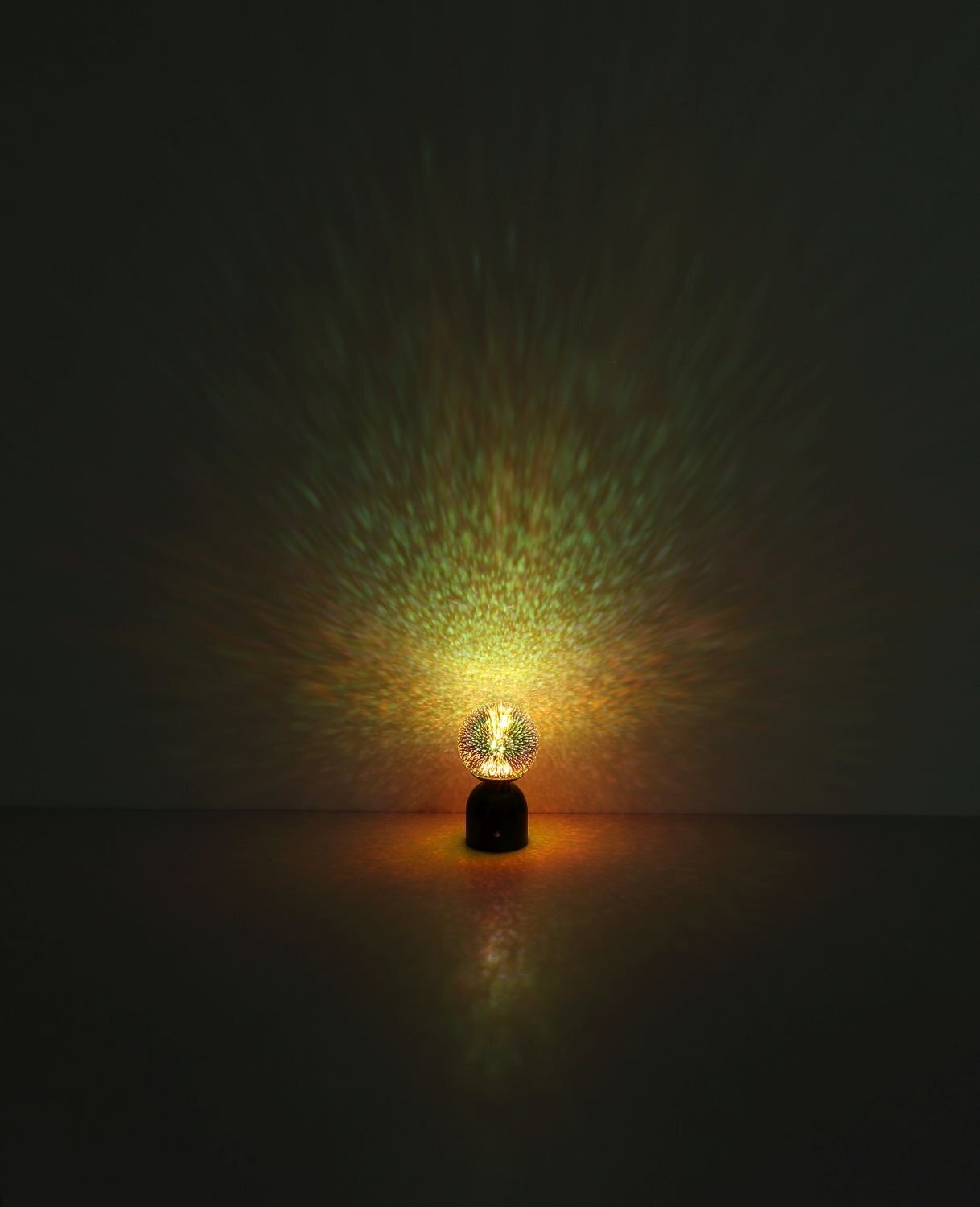 Na obrazie widoczna jest lampa stołowa emitująca kolorowe światło, tworzące wzory na ścianie. Lampa ma kulisty klosz i stoi na ciemnej podstawie.