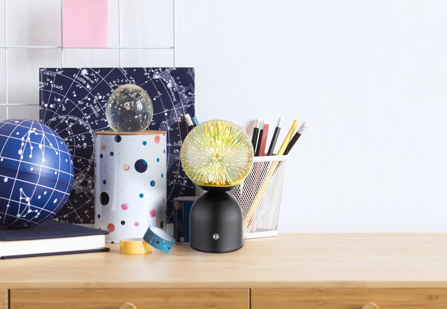 Na drewnianym blacie stoi stylowa lampa stołowa LED z czarną podstawą i żółtym, rozświetlonym kloszem. Obok znajdują się przybory do pisania w białym koszyku oraz dekoracje z motywem kosmosu.