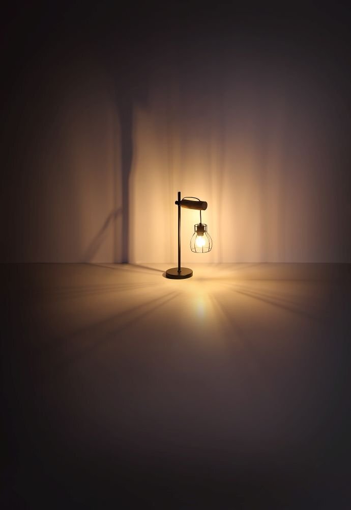 Na obrazie widoczna jest lampa biurkowa z czarnym stelażem i żarówką w kloszu z drutu, stojąca na gładkiej powierzchni. Światło lampy tworzy ciepłą poświatę na ścianie i podłodze.