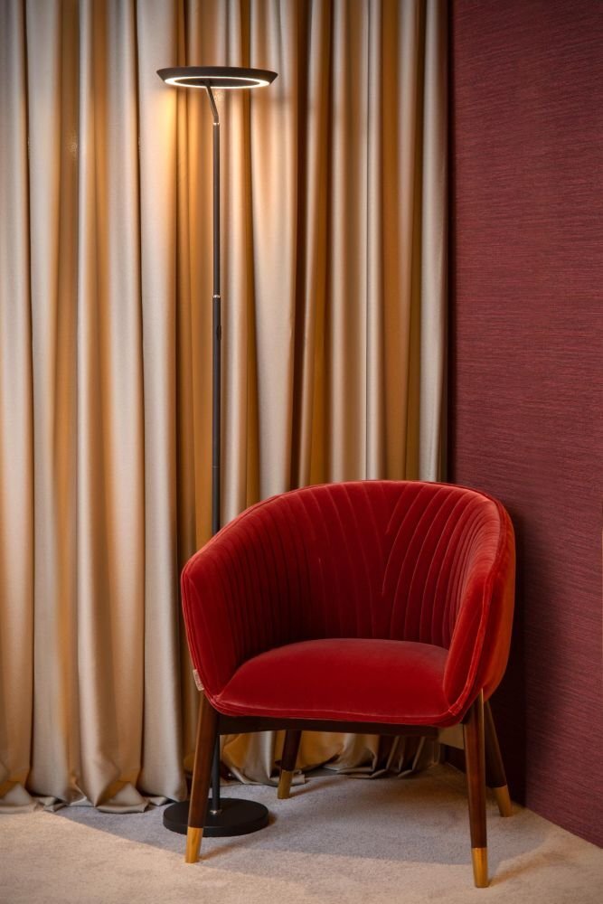 Na obrazie znajduje się lampa podłogowa LED o nowoczesnym designie, stojąca obok czerwonego, tapicerowanego fotela. W tle widoczne są zasłony w neutralnym kolorze oraz ściana w ciemnym odcieniu.
