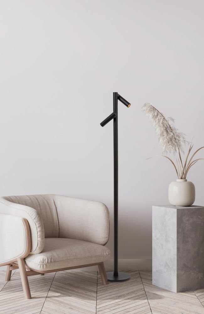 Na obrazie znajduje się czarna lampa podłogowa o nowoczesnym designie, stojąca obok beżowego fotela. Obok lampy znajduje się szara, betonowa kolumna z wazonem i suszonymi trawami.