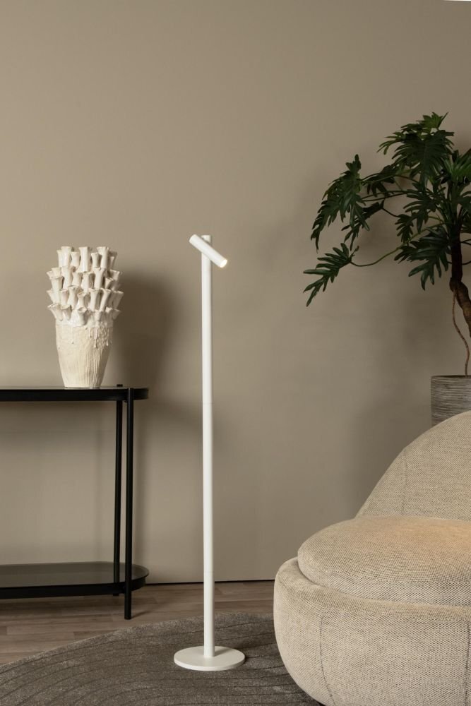 Na obrazie znajduje się biała, smukła lampa podłogowa z okrągłą podstawą, ustawiona w minimalistycznym wnętrzu. Obok lampy stoi czarna konsola z dekoracyjnym wazonem oraz roślina w doniczce.