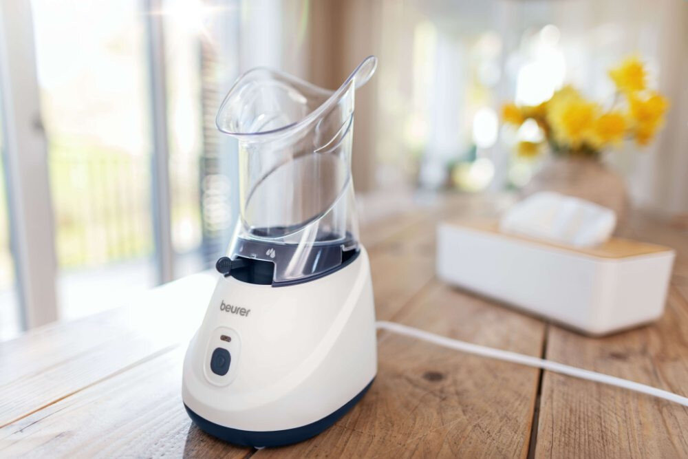 Na drewnianym stole stoi biały inhalator parowy z przezroczystą górną częścią. W tle widoczny jest rozmyty obraz żółtych kwiatów i pudełka z chusteczkami.