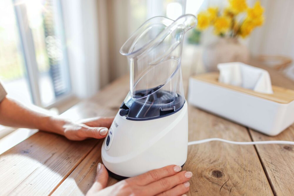 Na drewnianym stole znajduje się biały inhalator parowy z przezroczystą górną częścią. Obok widoczny jest biały pojemnik na chusteczki oraz żółte kwiaty w tle.