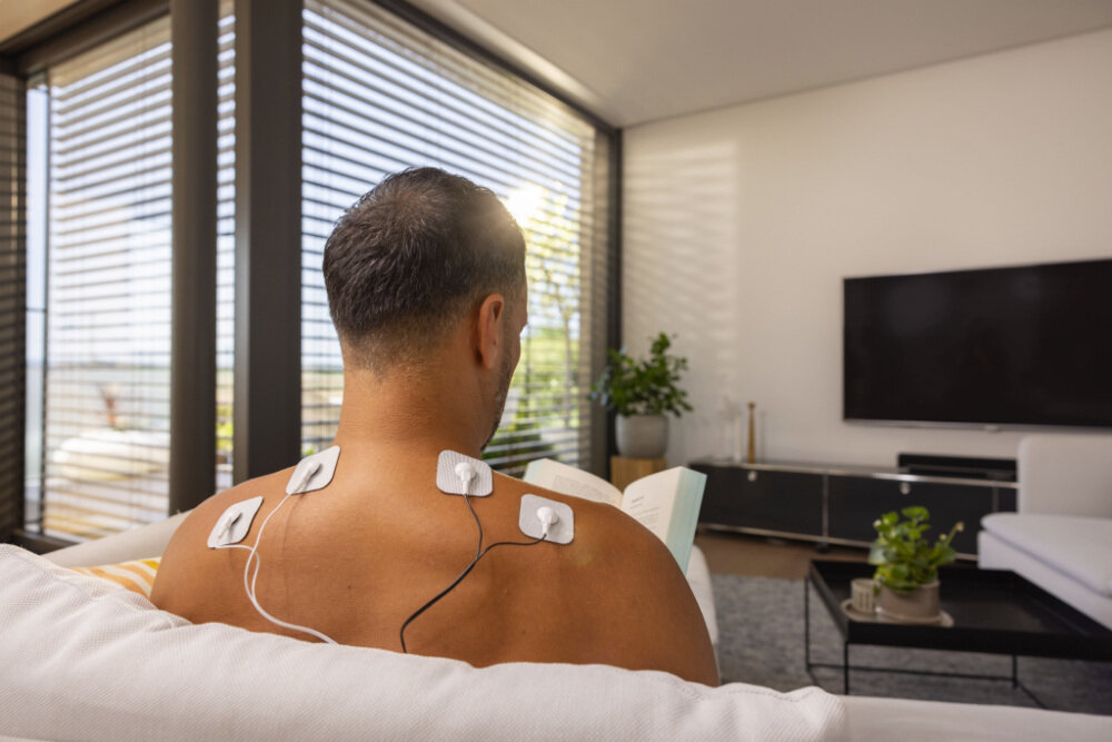 Osoba siedzi na kanapie z elektrodami przymocowanymi do pleców, trzymając książkę. W tle widać nowoczesne wnętrze z telewizorem i roślinami.