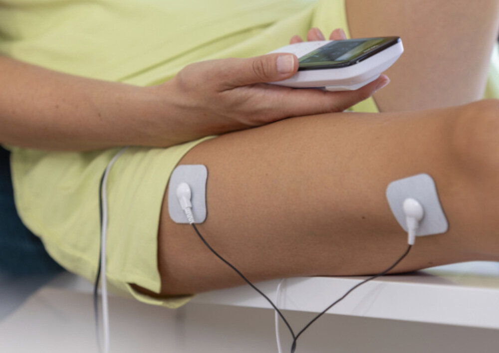 Osoba trzyma urządzenie elektroniczne, a na jej nodze znajdują się dwa elektrody przymocowane do skóry za pomocą przewodów.