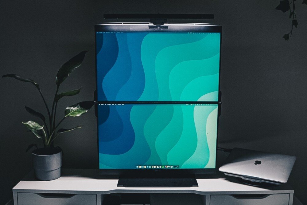 Monitor MOBILE PIXELS Geminos Dual Podwójny monitor ze scalonymi ekranami ustawiony pionowo na biurku, na którym wyświetla się abstrakcyjna falista grafika, obok stoi roślina doniczkowa