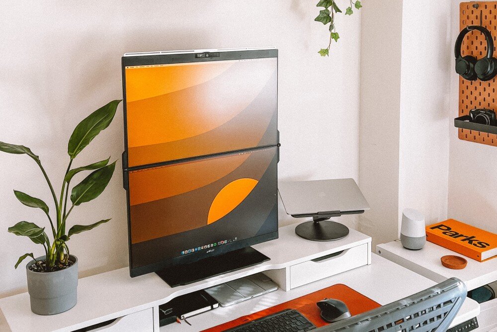 Monitor MOBILE PIXELS Geminos Dual Podwójny monitor pionowy stojący na jasnym biurku z otwartą przestrzenią, wyświetlający pomarańczowo-brązową grafikę, obok stoi zielona roślina