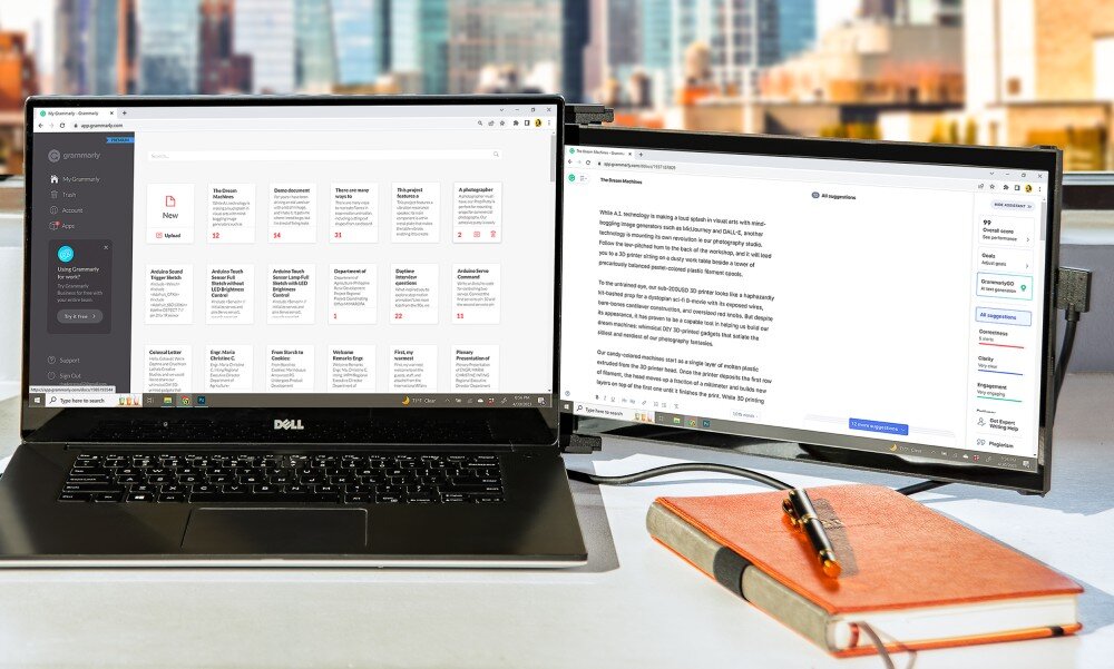 Monitor MOBILE PIXELS Duex Max DS Przenośny ekran przymocowany z prawej strony laptopa stojącego na biurku, w tle widoczne okno z widokiem na wieżowce, na pierwszym planie leży otwarty notes