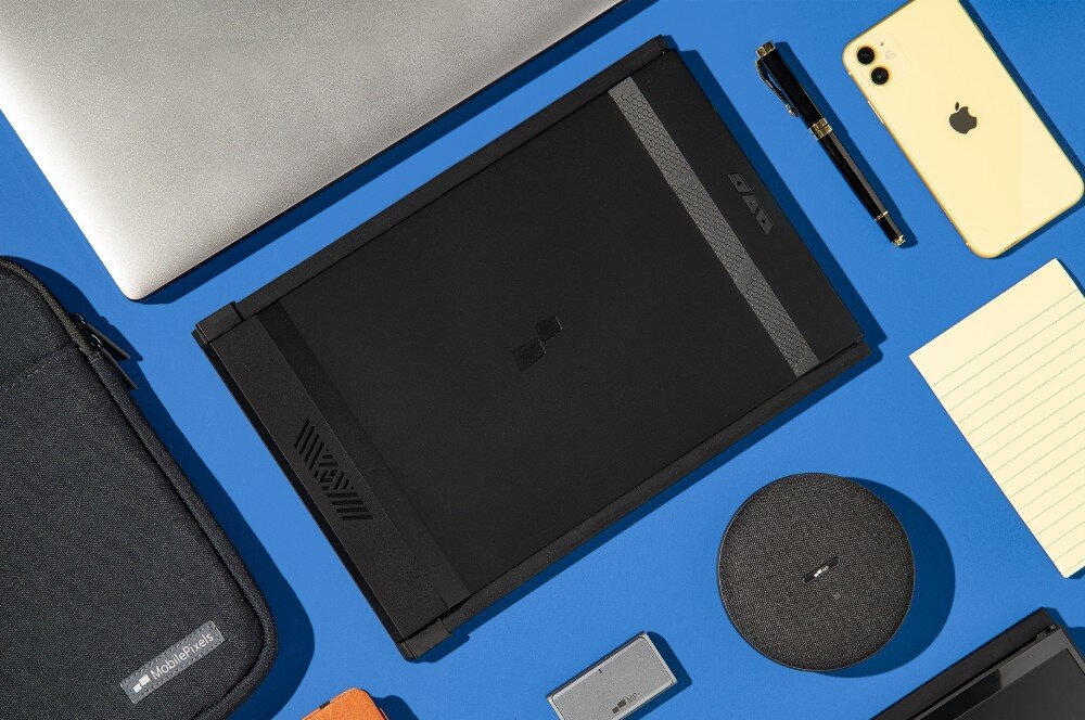 Monitor MOBILE PIXELS Duex Max DS Złożony ekran przenośny leżący na niebieskim tle w otoczeniu laptopa, etui, smartfona, długopisu, notesu oraz ładowarki indukcyjnej