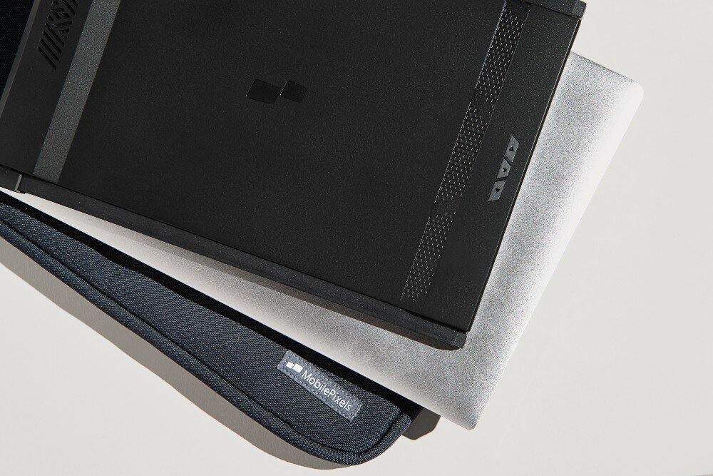 Monitor MOBILE PIXELS Duex Max DS Złożony monitor przenośny leżący na zamkniętym laptopie i materiałowym etui z logo MobilePixels, ujecie z góry na jasnym tle