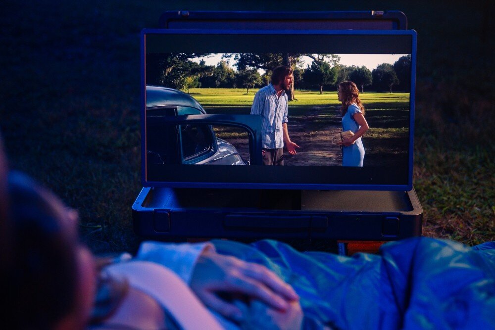 Monitor MOBILE PIXELS Pixcase Kobieta leżąca w śpiworze na trawie ogląda film wyświetlany na ekranie w otwartej walizce