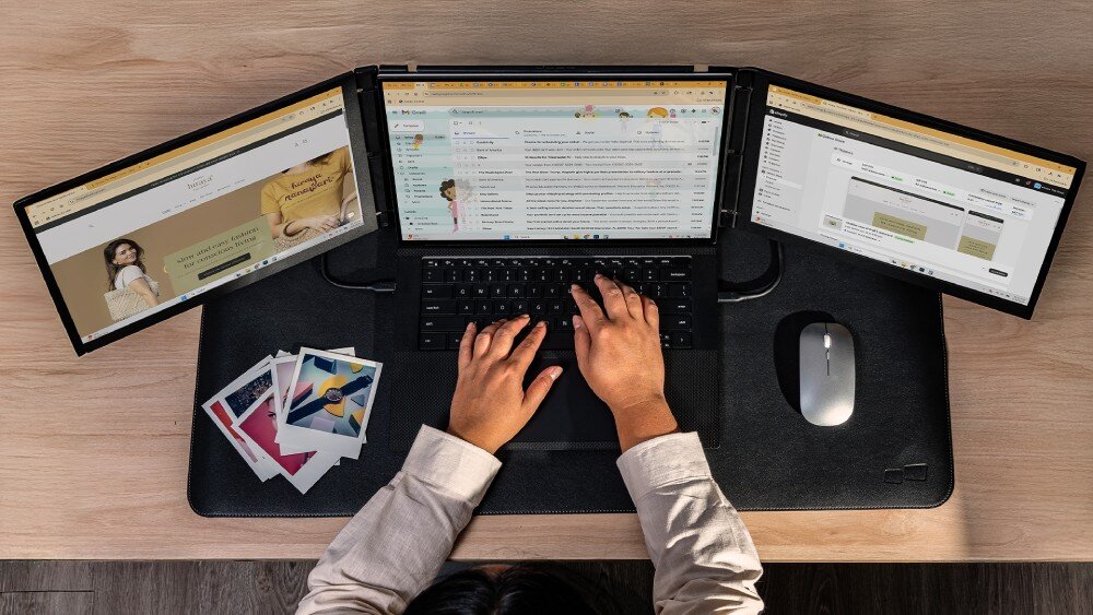 Monitor MOBILE PIXELS Trio 3 Pro Widok z góry na dłonie piszące na klawiaturze laptopa z dwoma bocznymi ekranami Przed urządzeniem leżą wydrukowane zdjęcia oraz myszka na podkładce