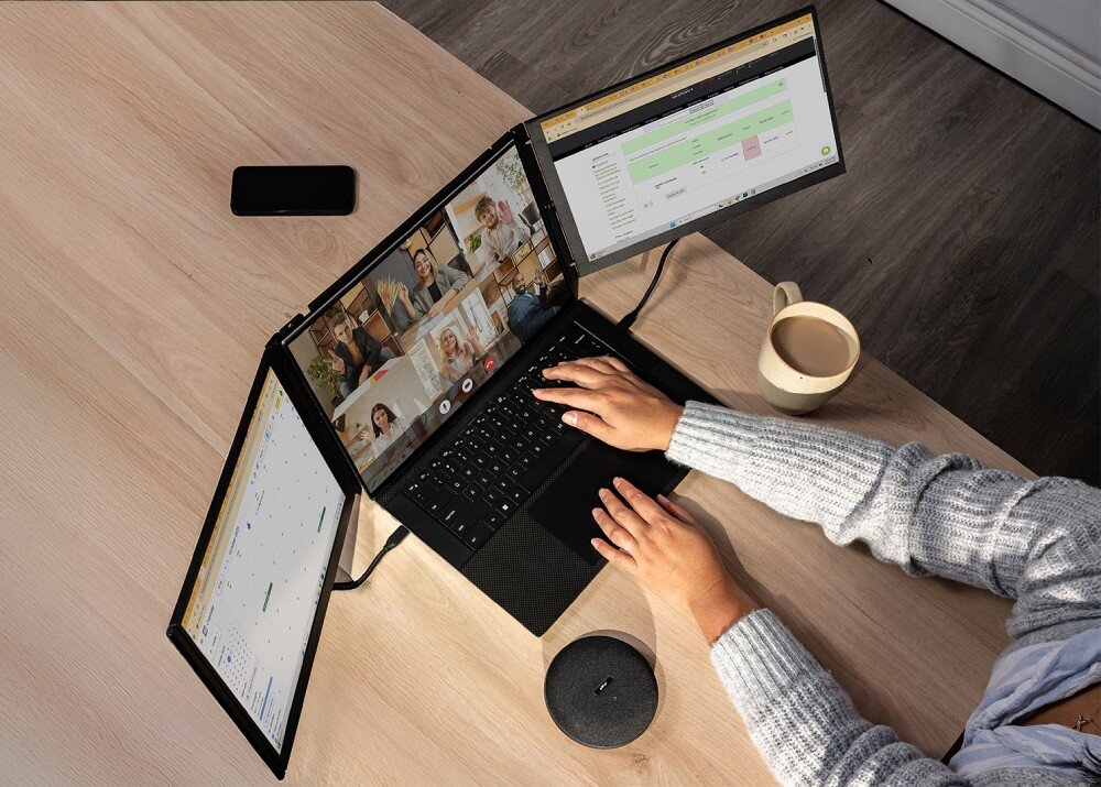 Monitor MOBILE PIXELS Trio 3 Pro Widok z góry na osobę pracującą na laptopie z dwoma dodatkowymi ekranami Na głównym wyświetlaczu trwa wideokonferencja, a obok stoją kubek i głośnik