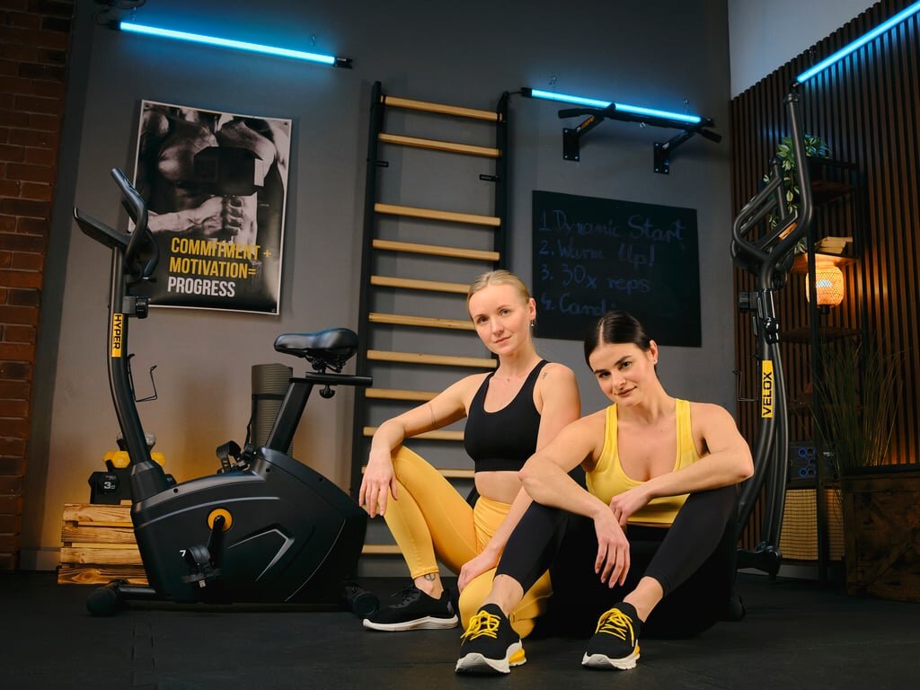 Na obrazie znajduje się rower stacjonarny magnetyczny z napisem \'HYPER\' oraz dwie osoby siedzące na podłodze w stroju sportowym. W tle widoczna jest drabinka gimnastyczna i plakat z napisem \'COMMITMENT = MOTIVATION = PROGRESS\'.
