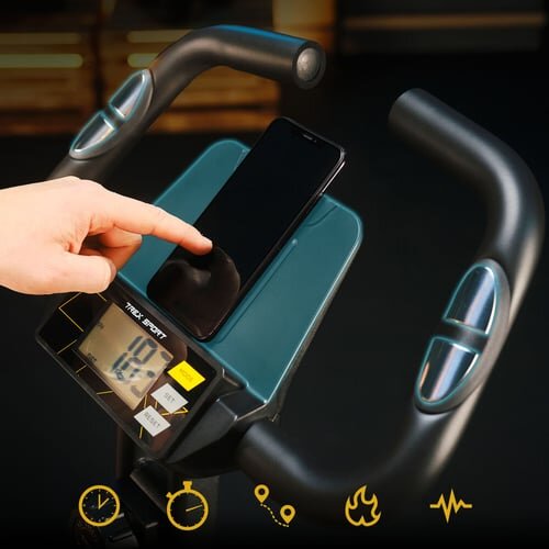 Osoba dotyka ekranu na panelu sterującym roweru stacjonarnego, na którym znajduje się telefon. Na panelu widoczny jest wyświetlacz z liczbą 18.0 oraz logo \'TREX SPORT\'.